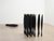 Around 20 identical tall, slender black sculptures are clustered together on a wooden gallery floor. Behind them, a smaller group of the same forms are stacked neatly on their side.
