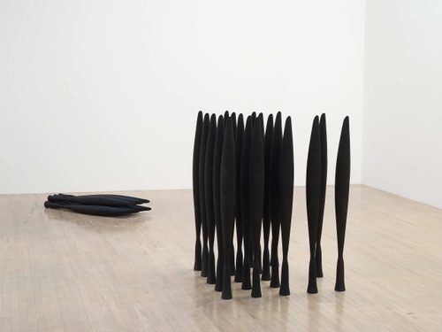 Around 20 identical tall, slender black sculptures are clustered together on a wooden gallery floor. Behind them, a smaller group of the same forms are stacked neatly on their side.