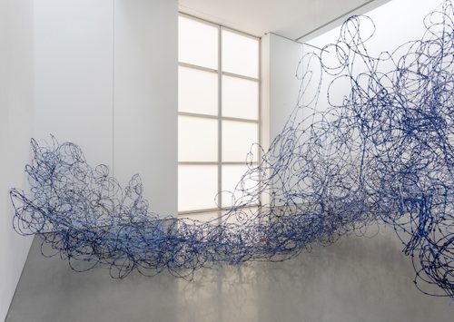 Photograph of the exhibition 'Lungiswa Gqunta: Sleep in Witness', showing the artist's sculpture 'Ntabamanzi' made from barbed wire and blue wool. The sculpture is designed to look like a wave breaking against the wall of the gallery.