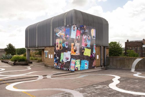 A billboard-sized aerial photo of people on a beach, with lots of colourful beach towels. It is mounted around two sides of the corner of a building.