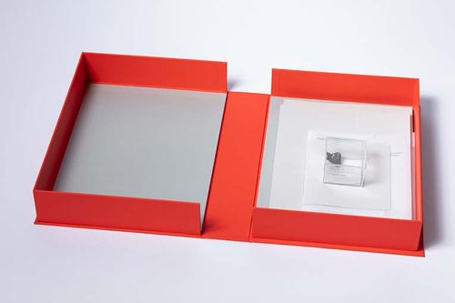 A red box, opened as a book would, containing some sheets of paper and a smaller plastic box with a stone inside.