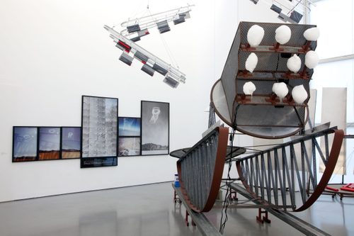 A large mechanical sled on rockers, mounted on rails. At the front is a stand, raised above the viewer, on which nine balls of candy floss are mounted. On the back wall are a series of framed photographs depicting landscapes with swirling vortices in the sky. Other abstract sculptural pieces hang from the ceiling, above and around the main 'sled' sculpture.