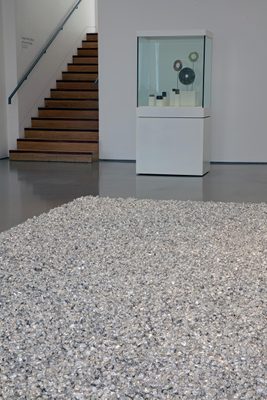 A square field of silver-wrapped sweets on a gallery floor, with a display cabinet behind holding rings of rock.