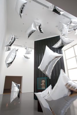 A display cabinet with the head of a damaged marble statue, surrounded by floating, pillow-shaped silver balloons.