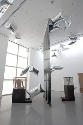 Two display cabinets holding damaged marble statues, separated by a vertical plastic sheet, and surrounded by floating, pillow-shaped silver balloons.