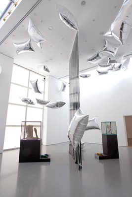 Two display cabinets holding damaged marble statues, separated by a vertical plastic sheet, and surrounded by floating, pillow-shaped silver balloons.