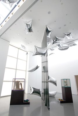 Two display cabinets holding damaged marble statues, separated by a vertical plastic sheet, and surrounded by floating, pillow-shaped silver balloons.