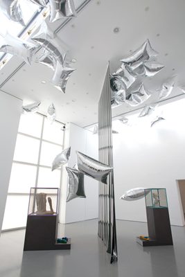 Two display cabinets holding damaged marble statues, separated by a vertical plastic sheet, and surrounded by floating, pillow-shaped silver balloons.