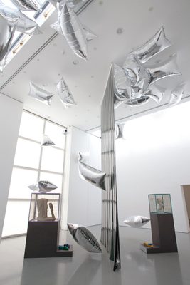Two display cabinets holding damaged marble statues, separated by a vertical plastic sheet, and surrounded by floating, pillow-shaped silver balloons.