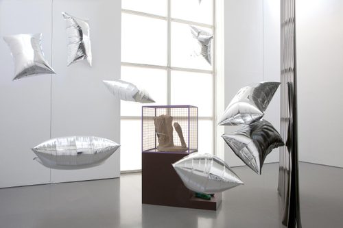 A display cabinet with the lower legs of a broken marble statue, surrounded by floating, pillow-shaped silver balloons.