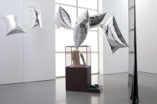 A display cabinet with the lower legs of a broken marble statue, surrounded by floating, pillow-shaped silver balloons.
