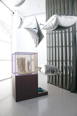 A display cabinet with the lower legs of a broken marble statue, surrounded by floating, pillow-shaped silver balloons.