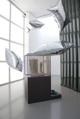A display cabinet with the lower legs of a broken marble statue, surrounded by floating, pillow-shaped silver balloons.