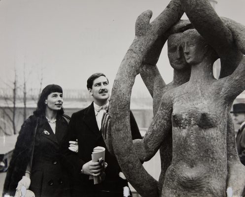 Black and white photo of a man and a woman looking at a sculpture of two people with branches twisting around them.