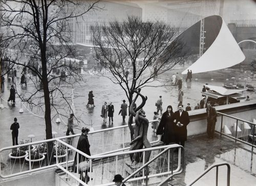 Black and white photo of a large outdoor exhibition space filled with people. In the foreground, a man and a woman are looking at a sculpture of two people with branches twisting around them.