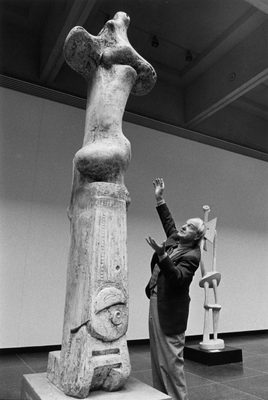 A black and white photo showing an older man, the sculptor Henry Moore, throwing his hands up in the air to demonstrate the size of the plaster sculpture he is stood next to.