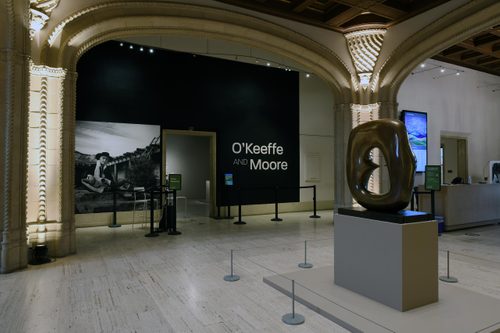 Entryway to an exhibition, with the title 'O'Keefe and Moore' in large white text on a black wall between two ornate pillars. There is a large black and white photo of a woman on the wall, and an oval bronze sculpture on a plinth.