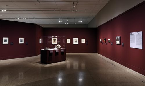 Wide shot of a gallery space, with several small paintings hung on a burgundy wall, and three small sculptures in a display case.