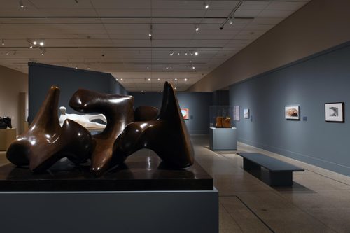 Close up view of a bronze sculpture by Henry Moore, made of three interlocking pieces similar to vertebrae. The negative space of the sculpture frames another behind it, this one of a reclining figure carved from white stone.