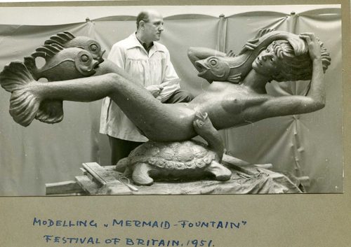 Black and white photo of an older man holding sculpting tools, stood behind a sculpture of a mermaid. The mermaid is reclining on a turtle, with two fish at her feet and another held close to her head.