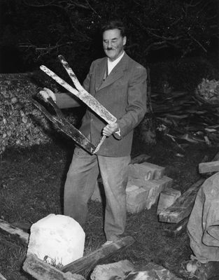 Black and white photo of an older man with a toothbrush moustache. He is standing outdoors, holding a large, bent piece of metal that will probably form part of a sculpture.