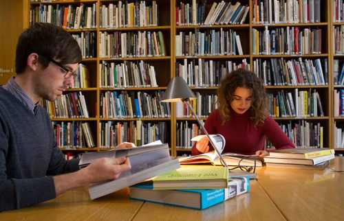 A man and woman are sat at a desk reading books about art.