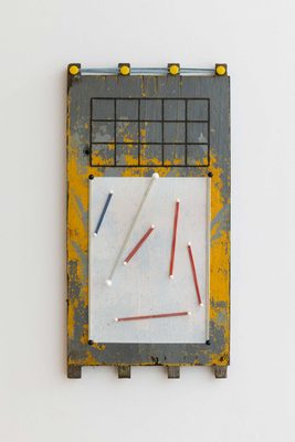 A grey-and-yellow-painted piece of wood, with a white rectangle on top, on which are several white pins with rubber bands stretched between them.