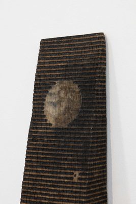 A wedge-shaped piece of wood with horizontal lines burned into it. There is a circle of negative space in the centre, where no lines have been burned.