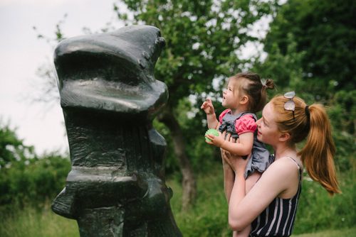 A teenager with red hair lifts a young toddler up to look more closely at a tall bronze sculpture