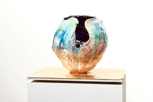A ceramic sculpture displayed on a plinth. The work is a vase type vessel with lustrous glaze, it has a void in one side showing a dark interior