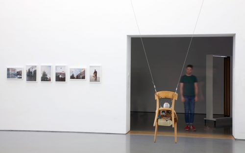A wooden chair is suspended in mid air on steel wires. Attached to the chair are the complicated apparatus or an exposed jet engine. On the wall next to the sculpture are six photos of the chair 'flying' as part of an event outside the Henry Moore Institute. A male visitor wearing a green t-shirt, blue jeans and red shoes looks at the sculpture from the adjoining gallery.