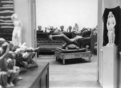 Black and white photo of an artist's studio, with a large mermaid sculpture at the centre and lots of smaller figurative sculptures and portrait busts around the room.