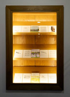 A large display cabinet set into a wall, with three rows of shelves, each displaying five illustrated pages , one with the title 'Virtuous Landscapes'.
