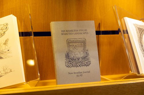 A book in a display case, with the title 'Ian Hamilton Finlay: Selected Landscapes - New Arcadian Journal 61/62'. There is an illustration of a simple tombstone with the word 'Fragile' on it.