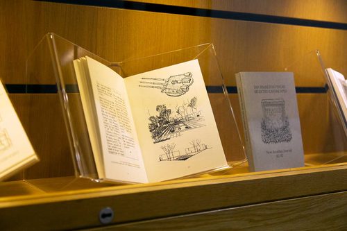 A book in a display case, opened at a page with an illustration of a tank turret with three gun barrels and two architectural drawings of a garden.