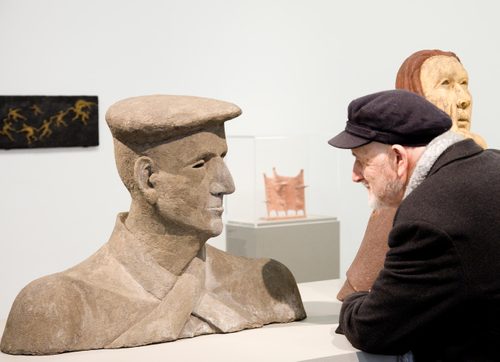 A man wearing a peaked hat looks at a large concrete portrait sculpture.