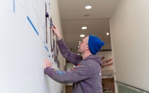 A man using a roller to paint large lettering on a white wall.